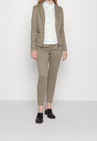 Blazer marron à motif pied-de-poule avec pantalon ajusté assorti, associé à une chemise blanche à col et à des mules noires à talons blocs. Texture lisse.
