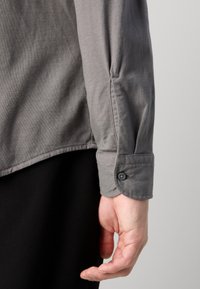 Grijze langemouwen shirt met een knoopmanchet, gemaakt van een gladde, textuurrijke stof en met een gebogen zoom, getoond tegen een zwarte achtergrond.