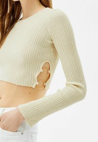 Crop top court côtelé beige clair à manches longues ; comprend deux anneaux dorés sur la découpe latérale. Texture douce et coupe ajustée.