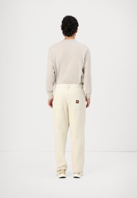 Bežs garām piedurknēm krekls, brīva piegriezuma baltas bikses ar aizmugurējiem kabatām un logo uzšuvumu, savienots ar gaišiem sporta apaviem.