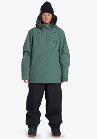 Zelená vodeodolná bunda so zipsom a kapucňou, skombinovaná so voľnými čiernymi nohavicami. Obe vyznačujú funkčným dizajnom pre vonkajšie aktivity.