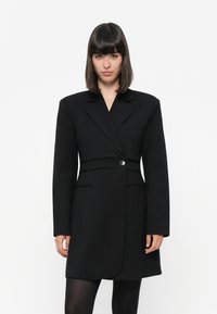 Svart skreddersydd blazer-kjole med lange ermer, enkel knappelukking, slagkrage, figurformet midje og lommer. Glatt tekstur.