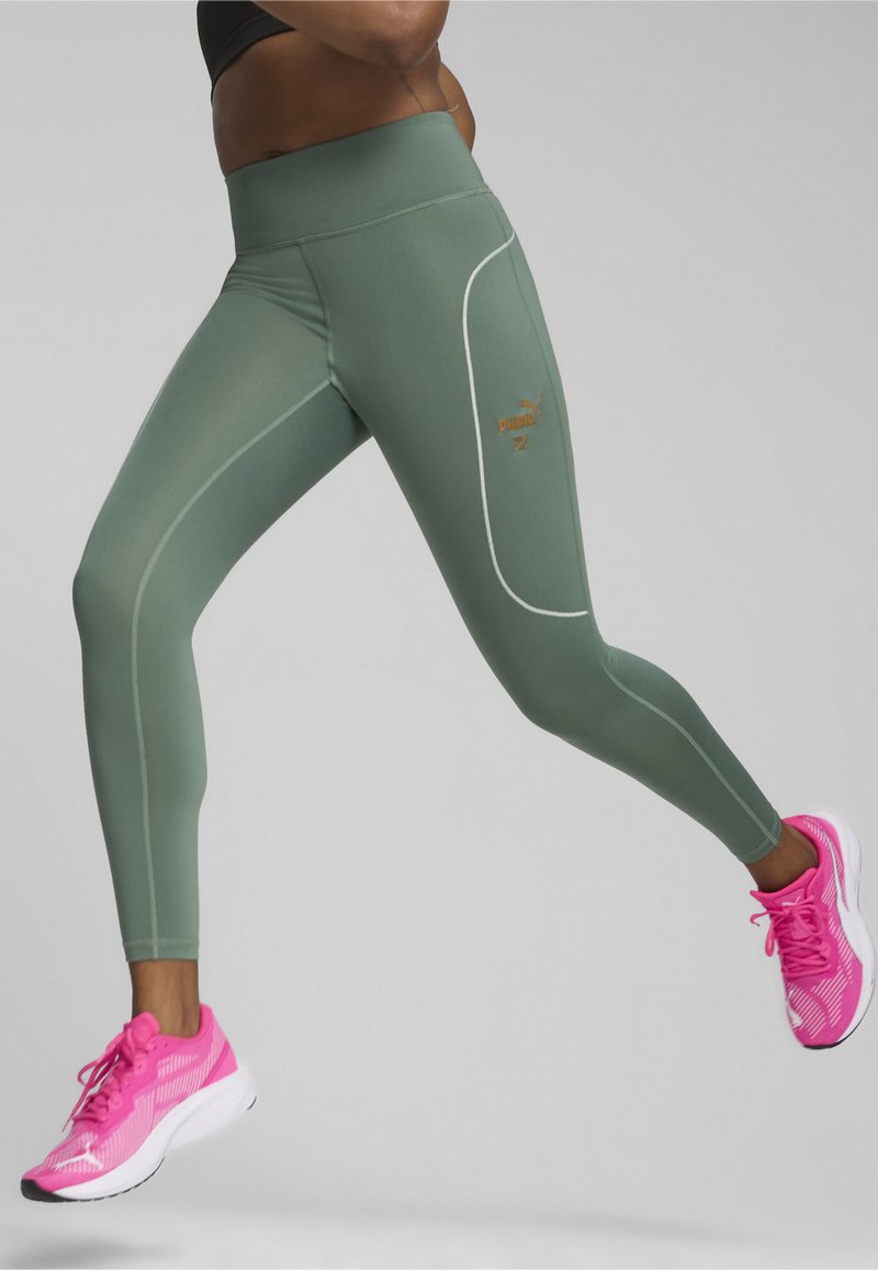 Leggings verdi a vita alta con cuciture bianche in evidenza e un piccolo logo dorato, abbinati a scarpe sportive rosa con suole bianche.