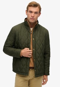 Hombre con chaqueta acolchada verde oliva sobre un suéter tan de media cremallera y pantalones beige, de pie frente a un fondo blanco.