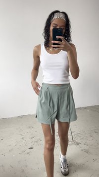 Hvit ermeløs topp, mintgrønne shorts med snøring, og svarte og hvite joggesko. Bakgrunnen er enkel med et nøytralt gulv.