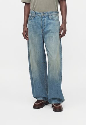 Personne portant un jean large bleu clair à taille haute et des chaussures en cuir marron foncé sans lacets.