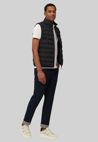 Schwarze Steppweste mit Steppdesign, getragen über einem weißen Hemd, kombiniert mit dunklen Jeans und hellen Sneakers. Einfacher, praktischer Stil.