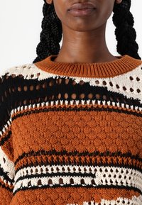 Neulotussa paidassa on pyöreä oranssi pääntie, jossa on mustia, valkoisia ja ruskeita raitoja, avoin neulos ja väljä istuvuus.