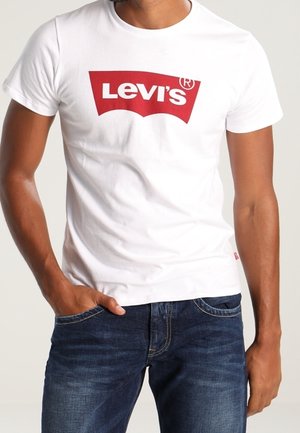 Biała bawełniana koszulka z krótkim rękawem z czerwonym logo Levi's z przodu, w zestawieniu z niebieskimi dżinsami. Prosty design, okrągły dekolt.