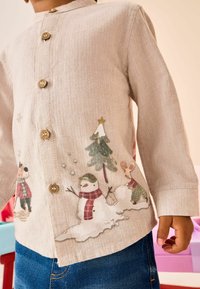 Chemise boutonnée beige clair avec une texture en chevron, présentant des illustrations colorées de bonhommes de neige et d'arbres, accentuée par quatre boutons en bois naturel.