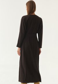 Langes, dunkelbraunes Kleid mit langen Ärmeln und tailliertem Schnitt. Glatter, strukturierter Stoff mit einer schmalen Silhouette und seitlichen Schlitzen.