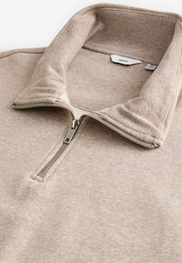 Beige Kapuzenpullover mit einem weichen, strukturierten Stoff, einem hohen Kragen und einem silbernen Reißverschluss. Das Etikett trägt die Aufschrift "NEXT". Keine sichtbaren Muster oder Akzente.
