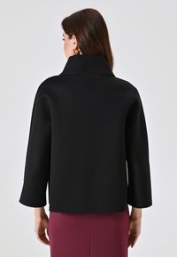 Chaqueta negra corta con mangas anchas y un cuello alto. Textura suave, diseño minimalista, combinada con una falda morada, vista desde atrás.