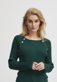 Maglione verde a coste con scollo rotondo e dettagli di bottoni argentati sulle spalle. Maniche lunghe e tessuto testurizzato.