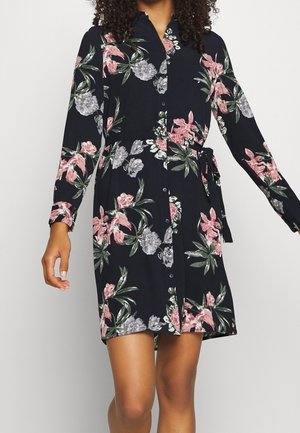 Woman wearing a black floral button-up dress with long sleeves and a waist tie, showing pink, gray, and green flower patterns.
