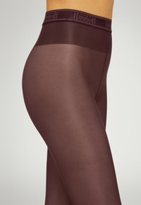 Collant opachi bordeaux con una texture liscia, dotati di un elastico in vita con logo. I collant avvolgono visibilmente le curve delle gambe.