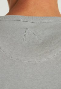 Pilkas medvilninis marškinėlis su apvalia iškirpte, ant nugaros turi subtilų siuvinėjimo detalę trikampio forma. Lygi tekstūra.