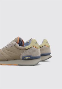 Baskets beiges avec une tige en daim et mesh, accents orange et jaune, col rembourré, logo bleu sur le talon, semelle en caoutchouc grise.