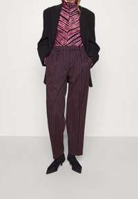 Černý blazer přes růžový halenkový top se zebrovaným vzorem a vysokým límcem, doplněný širokými kalhotami s vysokým pasem a pruhy v tmavých tónech a černými podpatky.