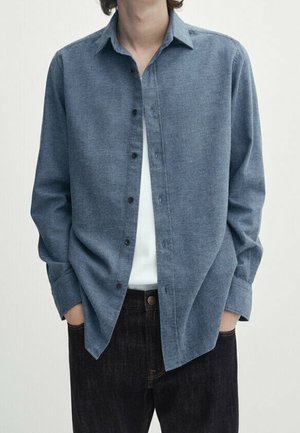 Person iført en åben blå langærmet skjorte over en hvid T-shirt med hænderne i lommerne på mørke jeans.