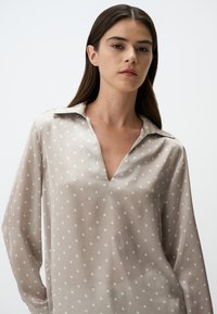 Blusa beige chiaro con pois bianchi, scollatura a V, maniche lunghe e una consistenza liscia e setosa. Vestibilità leggermente ampia per il comfort.