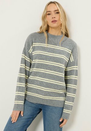 Frau trägt einen grauen Strickpullover mit blassgelben horizontalen Streifen und blaue Jeans, steht vor einer hellen Wand.