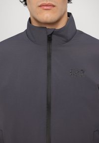 Grå zip-up jakke med høy krage, laget av glatt, lett materiale. Har en liten svart EA7-logo foran.
