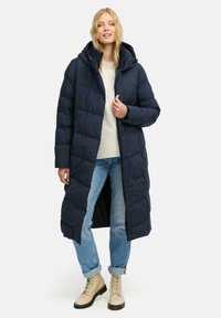 Lång marinblå pufferjacka med huva, med knappstängning, kviltat mönster och sidofodringar. Kombinerad med ljusa jeans och beige stövlar.