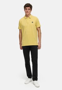 Gul pikétröja i bomull, med krage och en liten svart logotyp. Tillsammans med svarta byxor och vita sneakers.