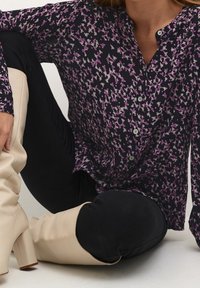 Langærmet bluse i mørklilla med blomsterprint i pink og hvid, med knaplukning foran og en afslappet pasform. Sorte bukser og beige knæhøje støvler.