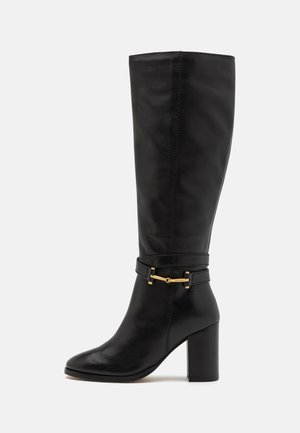 Bottes en cuir noir à hauteur de genou avec un talon épais, dotées d'une boucle dorée sur une sangle noire autour de la cheville. Texture lisse.