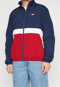 Granatowo-czerwona i biała kurtka wiatrówka z zamkiem błyskawicznym z przodu, wysokim kołnierzem, elastycznymi mankietami oraz markowym logo na piersi.