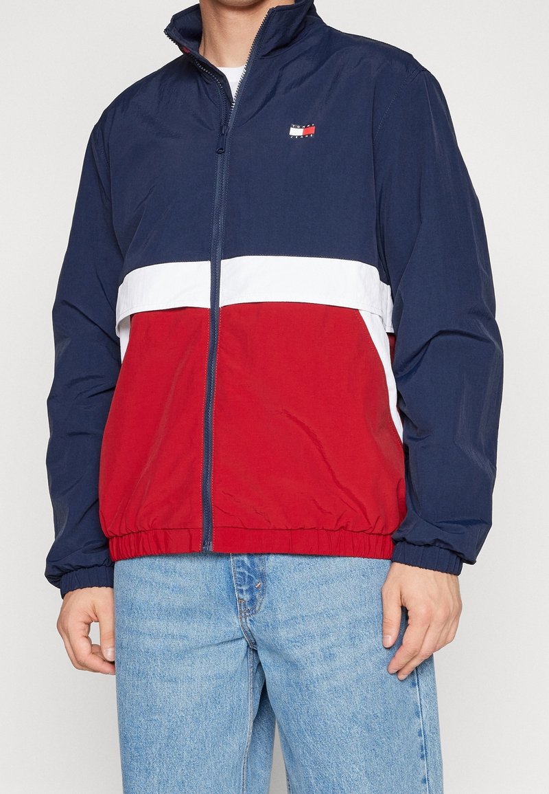 Granatowo-czerwona i biała kurtka wiatrówka z zamkiem błyskawicznym z przodu, wysokim kołnierzem, elastycznymi mankietami oraz markowym logo na piersi.