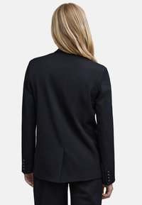Blonde vrouw in een zwarte op maat gemaakte blazer en bijpassende broek, gezien van achteren tegen een effen achtergrond.