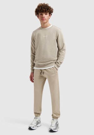 Jonge man met krullend haar, gekleed in een beige sweatshirt en bijpassende joggingsbroek, wit T-shirt eronder en zilveren hardloopschoenen, staand ontspannen.