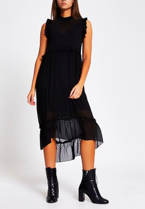 Woman wearing a sleeveless black midi dress with ruffles and sheer polka dot fabric, paired with black high-heeled lace-up ankle boots.