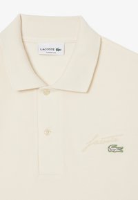 Lacoste klassisk passform polo i krämfärg med en texturerad tyg, treknapps låsning, broderad logotyp och subtil brandingdetalj på bröstet.