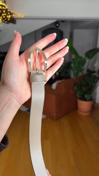Beiger Ledergürtel mit einer goldfarbenen Metallschnalle, zeichnet sich durch eine glatte Textur und ein schlichtes Design aus. Eine Hand hält den Gürtel.