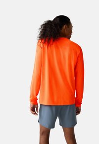 Person med mørkt krøllet hår i hestehale iført en lys orange langærmet skjorte og grå shorts, vendt væk på en ensfarvet baggrund.