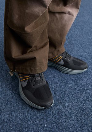 Czarne buty sportowe z siateczkowymi wstawkami, pomarańczowymi akcentami i warstwową szaro-srebrną podeszwą, w zestawieniu z luźnymi, brązowymi spodniami z materiału.