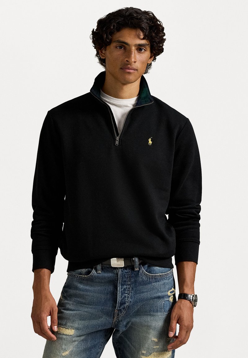 Ung mand med krøllet hår iført en sort kvart-zip trøje med et gult logo, hvidt underskorte, slidte blå jeans og et armbåndsur.