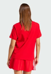 Rød t-shirt med korte ærmer, lavet af bomuld. Har hvide striber på skuldrene og en lagdelt, udskåret kant forneden.