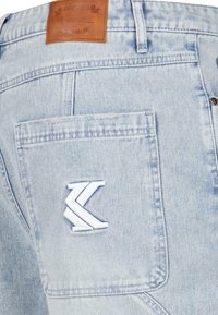 Jeans de mezclilla de color azul claro con un parche de cuero en la parte trasera y un logo bordado en blanco en el bolsillo derecho. Diseño clásico de cinco bolsillos.