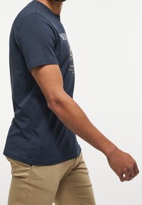 Námořnický modrý tričko s krátkým rukávem a kulatým výstřihem, s grafickým potiskem na přední straně. Nosené s světle khaki kalhotami.