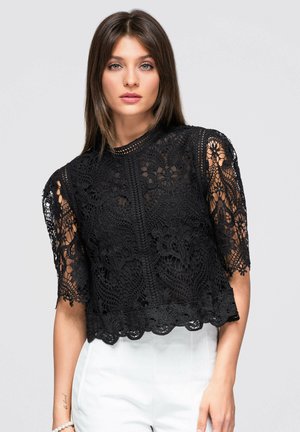 Femme aux cheveux bruns lisses portant un haut noir en dentelle à manches courtes et un pantalon blanc taille haute sur un fond clair uni.