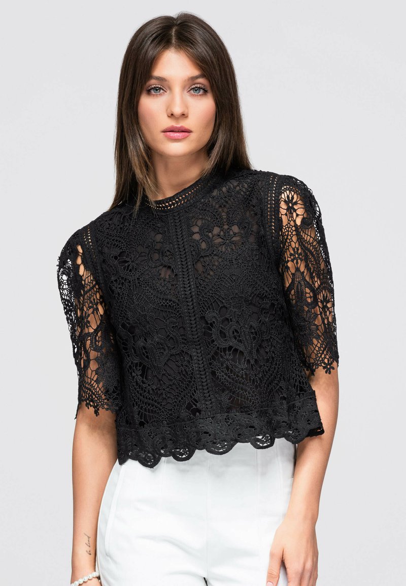 Femme aux cheveux bruns lisses portant un haut noir en dentelle à manches courtes et un pantalon blanc taille haute sur un fond clair uni.