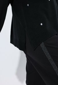 Maglione nero lavorato a maglia con gemme decorative trasparenti, spacchi laterali e orlo a coste, abbinato a pantaloni neri con una striscia verticale lucida.