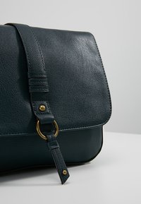 Mörkgrön lädercrossbody-väska med klaffstängning, som har en guldfärgad accentring och texturerad yta på kroppen och axelremmen.