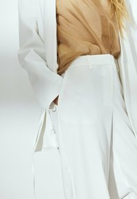 Blazer blanc ajusté avec un pantalon assorti, accompagné d'une blouse translucide beige. Texture lisse, lignes épurées, et un détail de ceinture sur le pantalon.