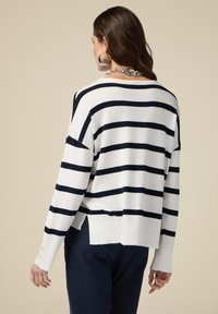 Maglione bianco con righe orizzontali blu marino, vestibilità rilassata, aperture laterali e bottoni decorativi sulle maniche. Texture liscia, design informale.